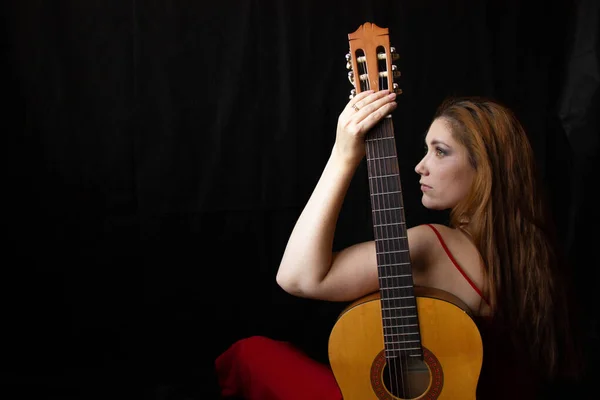 Menina em um vestido vermelho senta-se no chão perto da guitarra em um fundo preto — Fotografia de Stock