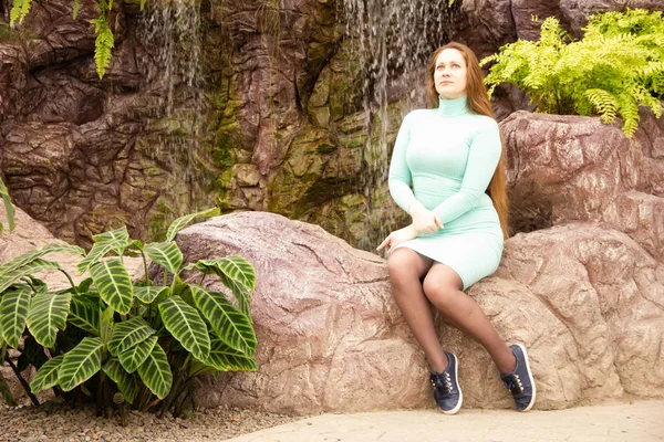 Krásná mladá dívka sedí na kamenech v blízkosti umělé nádrže a vodopádu — Stock fotografie
