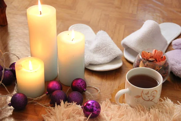 Brandende witte kaarsen staan op de vloer, naast ligt een pluizige ruit, twee paar pantoffels en een mok thee — Stockfoto