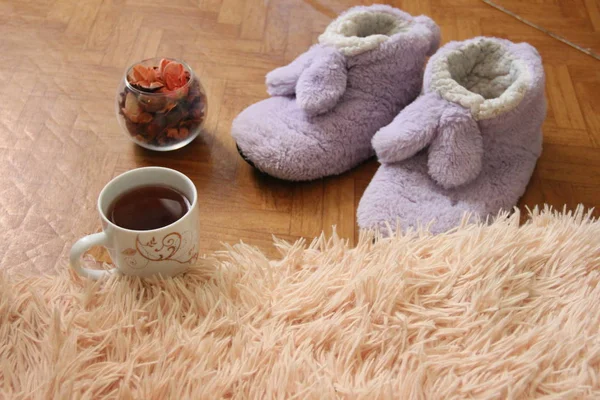 a pair of slippers on the floor next to a mug of tea and a fluffy plaid