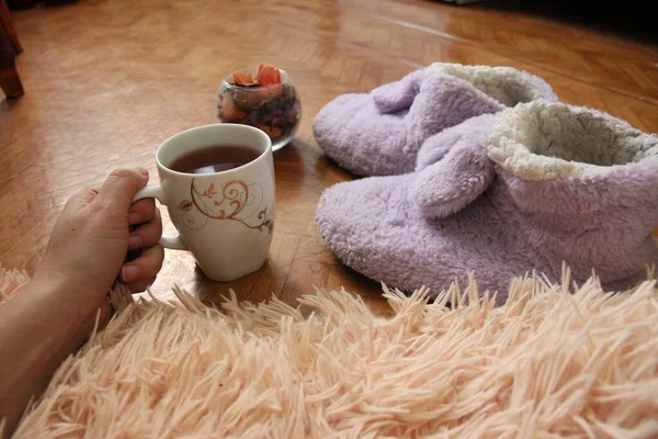 Hand houdt een mok thee. slippers op de vloersteun en naast de pluizige ruitjes — Stockfoto