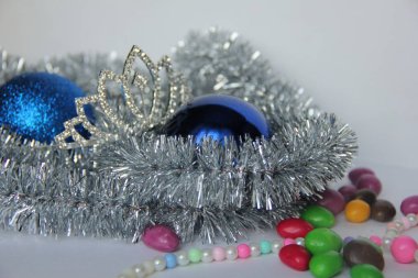 Taç, mavi Noel toplarının yanında gümüş bir süslemenin üzerinde duruyor. Beyaz arka planda çok renkli boncuklar ve çok renkli şekerler var..