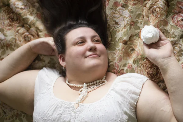 Beyaz elbiseli şişman seksi bir kadın yatağa uzanıyor ve elinde iki pasta tutuyor..
