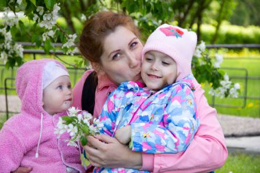 Annem çiçek açan bir elma ağacının gölgesinde çimlerin üzerinde oturur. Oynuyor ve kızlarına sarılıyor..
