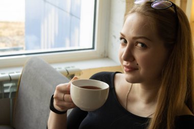 Siyah elbiseli güzel seksi kız kanepenin penceresinde oturur ve beyaz bardaktan sıcak su içer. 