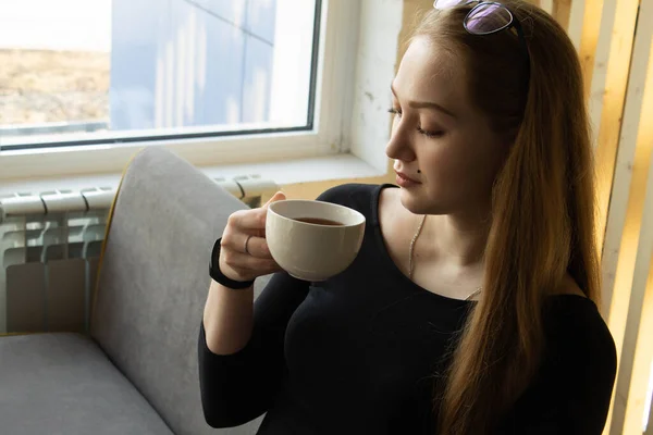 Beautiful sexy girl in black dress sits by the window on the couch and drinks from a white cup hot drink — Stock Photo, Image