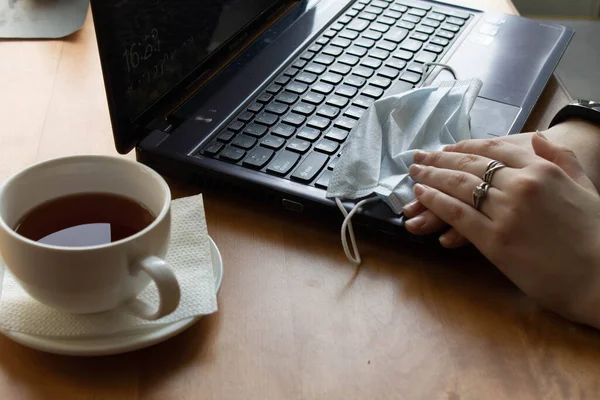 Sobre la mesa se encuentra el portátil, junto a una taza de té. las manos yacen sobre una máscara médica en el teclado — Foto de Stock