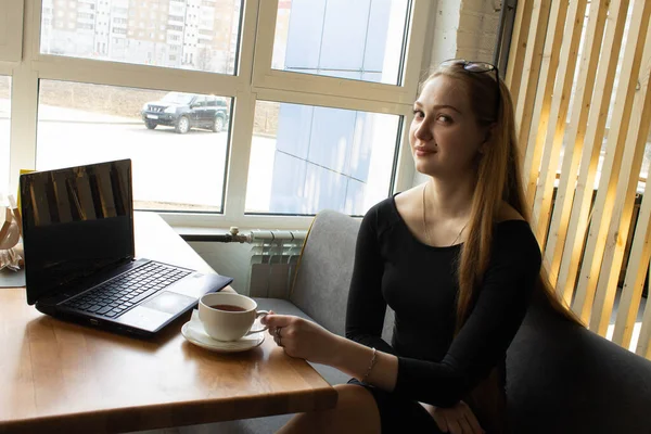 A beautiful girl in a black dress sits at a table by the window. Break work or computer training