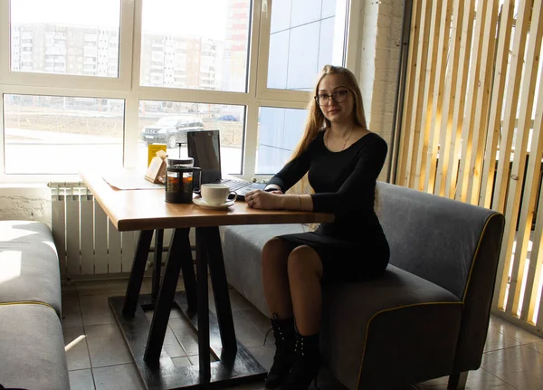 검은 옷을 입고 안경을 쓴 아름다운 섹시 한 소녀가 식탁에 앉아 컴퓨터로 일합니다. 거리 학습. 외딴곳에서의 활동 — 스톡 사진