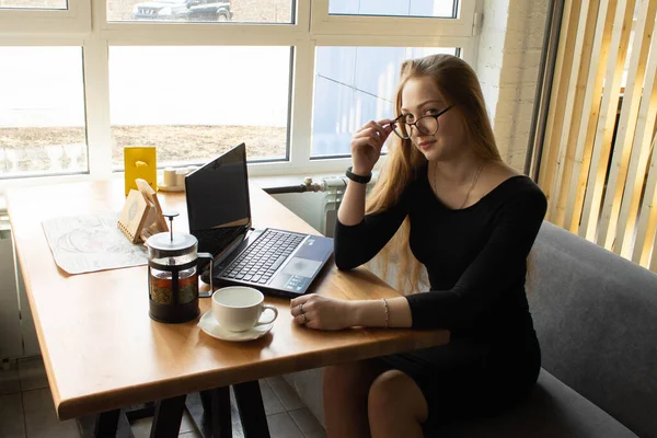 Красивая сексуальная девушка в черном платье и очках сидит за столом и работает за компьютером. Дистанционное обучение. Удаленная работа — стоковое фото
