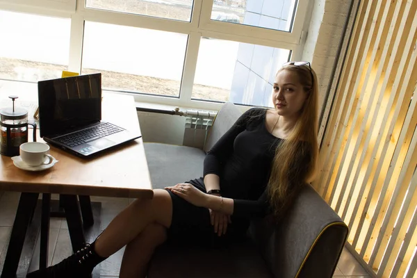 A beautiful girl in a black dress sits at a table by the window. Break work or computer training