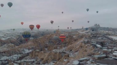 Kapadokya, Türkiye 'de kış mevsiminde karla kaplı perili bacaların üzerinden uçan bir sürü sıcak hava balonunun 4k hava manzarası.