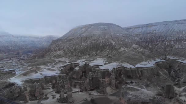 トルコのカッパドキアで雪の冬の風景とパサバグ渓谷の大きな妖精の煙突の中の小さな洞窟の窓に一人で立っている女性の空中叙事詩の景色 — ストック動画