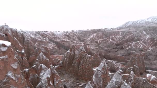 トルコのカッパドキアにあるImaginary Valleyの岩の層に囲まれた妖精の煙突と洞窟の家 カッパドキアのデバントバレーで雪で覆われたトゥファ層 — ストック動画