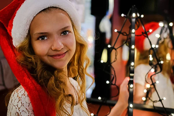 Merry Christmas and happy holidays. A cute blonde girl in a Santa Claus hat stands next to a mirror. Girl enjoys the holiday on a dark background.