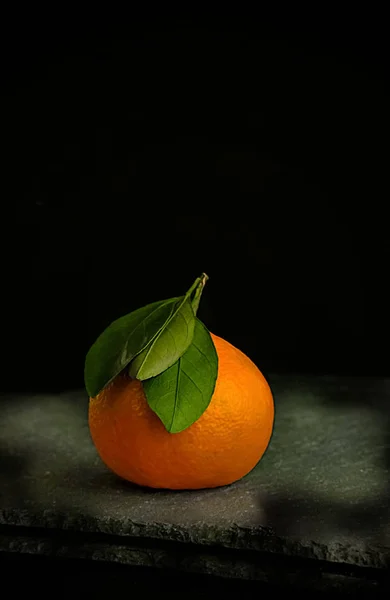 Ripe juicy tangerine with a green leaf on a black background, lit by the rays of the sun — Stock Photo, Image