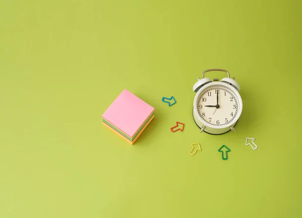 Ρολόι με ένα ξυπνητήρι σε λευκό φόντο βρίσκεται σε χρωματιστό χαρτί φύλλα από πράσινο, μπλε, ροζ, κίτρινο και βατόμουρο χρώμα για σημειώσεις — Φωτογραφία Αρχείου