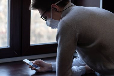Yeniden kullanılabilir tıbbi maskeli genç bir adam kendini koronavirüsten nasıl koruyacağına dair bilgi almak için telefon ekranına bakar.
