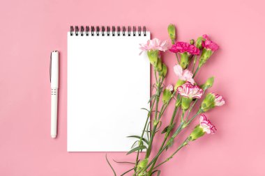 Renkli buket pembe karanfil çiçekleri, beyaz defter, pembe arka planda kalem Üst görünümde düz tatil kartı