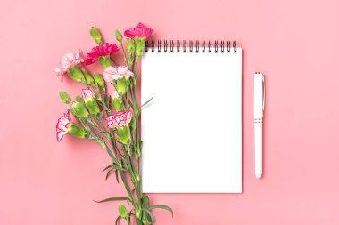 Farklı pembe karanfil çiçeklerinden oluşan renkli buket, beyaz defter, pembe arka planda bir kalem. Üst tarafta düz bir tatil kartı var. 8 Mart, Sevgililer Günü, Anneler Günü konsepti.