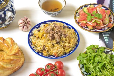 Ulusal Özbek pilavı et, domates salatası, salatalık, geleneksel desenli soğan, kişniş, kiraz domatesleri, sarımsak, ekmek tortilla - beyaz ahşap masa manzaralı.