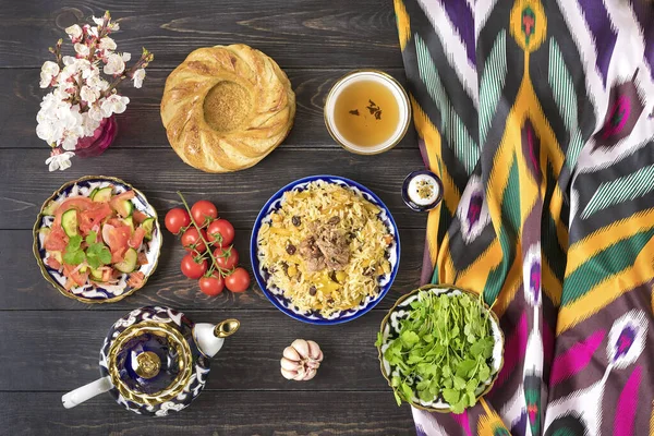 Ulusal Özbek pilavı et, domates salatası, salatalık, geleneksel desenli soğan, kişniş, kiraz domatesleri, sarımsak, ekmek tortilla - koyu ahşap masa manzaralı.
