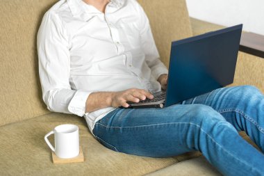  Beyaz gömlekli ve kot pantolonlu genç adam dizüstü bilgisayarda çalışıyor, ev bilgisayarında yazıyor rahat bej koltukta oturuyor evde oturuyor, online çalışıyor, uzaktan öğrenme konsepti geliştiriyor. 
