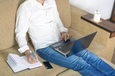  Beyaz gömlekli ve kot pantolonlu genç adam dizüstü bilgisayarda çalışıyor, ev bilgisayarında yazıyor rahat bej koltukta oturuyor evde oturuyor, online çalışıyor, uzaktan öğrenme konsepti geliştiriyor. 