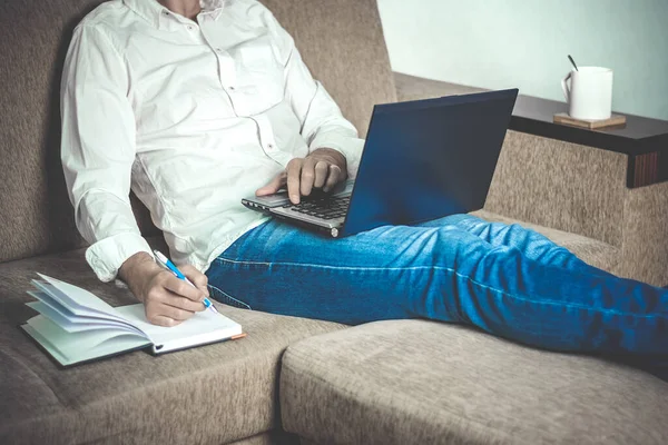  Beyaz gömlekli ve kot pantolonlu genç adam dizüstü bilgisayarda çalışıyor, ev bilgisayarında yazıyor rahat bej koltukta oturuyor evde oturuyor, online çalışıyor, uzaktan öğrenme konsepti geliştiriyor. 