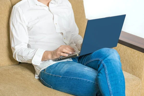 Hombre joven en camisa blanca y vaqueros azules que trabajan en el ordenador portátil, escribe en el cuaderno, mirando en la pantalla blanca en la oficina en casa sentado en el sofá beige acogedor Quédese en casa, trabajo en línea, aprendizaje a distancia — Foto de Stock