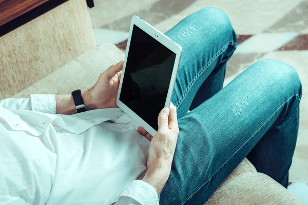 Joven con camisa blanca y vaqueros azules trabajando y mirando en la tableta con pantalla negra en la oficina en el acogedor sofá beige Quédese en casa, trabajo en línea, concepto de aprendizaje a distancia — Foto de Stock