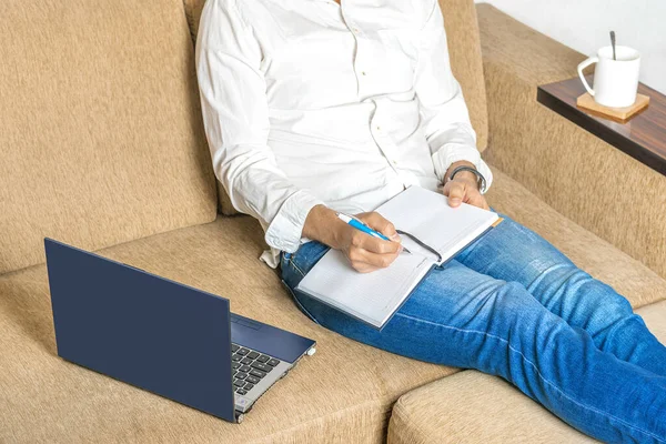 Hombre joven en camisa blanca y vaqueros azules que trabajan en el ordenador portátil, escribe en el cuaderno en la oficina en casa sentado en el sofá de color beige acogedor — Foto de Stock