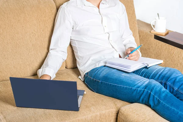 Hombre joven en camisa blanca y vaqueros azules que trabajan en el ordenador portátil, escribe en el cuaderno en la oficina en casa sentado en el sofá de color beige acogedor — Foto de Stock