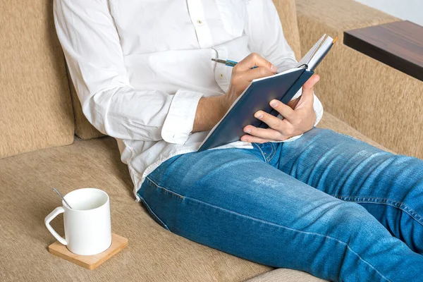 Cup of tea, young man in white shirt and blue jeans working at home, writes in notebook in home office sitting on cozy beige sofa Stay at home, stay safe. Online work, distance learning concept