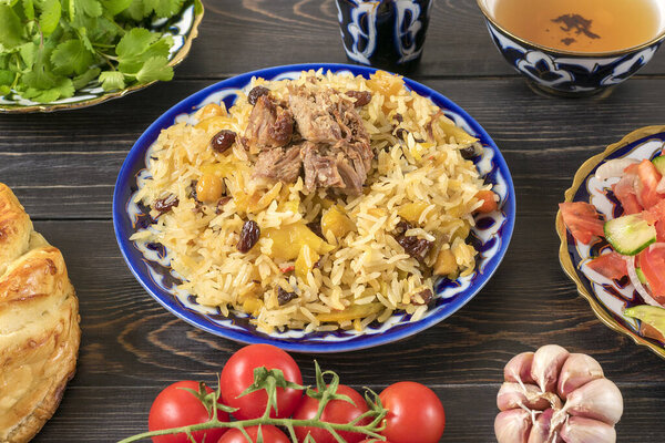 National Uzbek pilaf with meat, achichuk salad of tomato, cucumber, onion in plate with traditional pattern, cilantro, cherry tomatoes, garlicbread tortilla - patir on dark wooden table Top view.