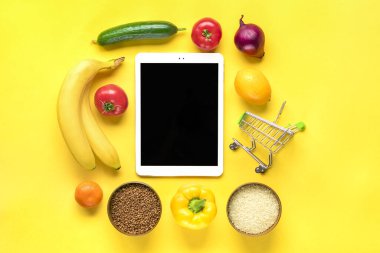 farklı sağlıklı yiyecekler - karabuğday, pirinç, sarı dolmalık biber, domates, muz, marul, yeşil, salatalık, soğan, sarı arka planda siyah ekranlı tablet düz görünüm düz çizgi üzerinde alışveriş