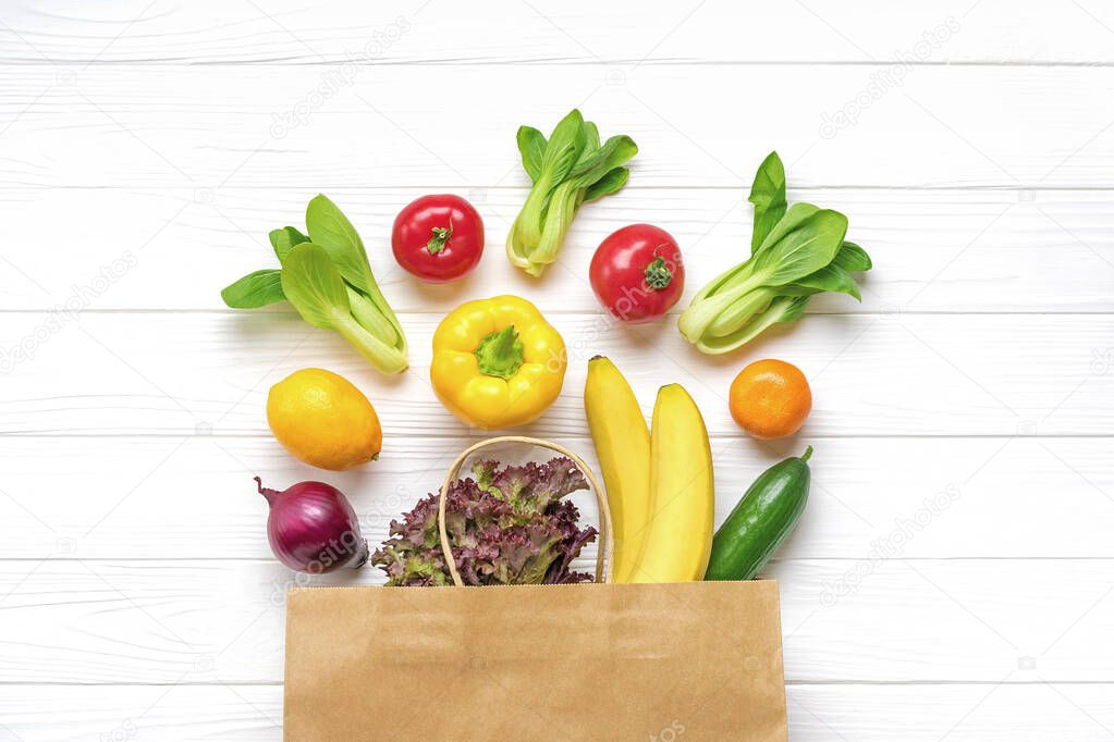 Full eco paper bag of different health food - yellow bell pepper, tomatoes, bananas, lettuce, green, cucumber, onions on white wooden background Top view Flat lay Grocery shopping.