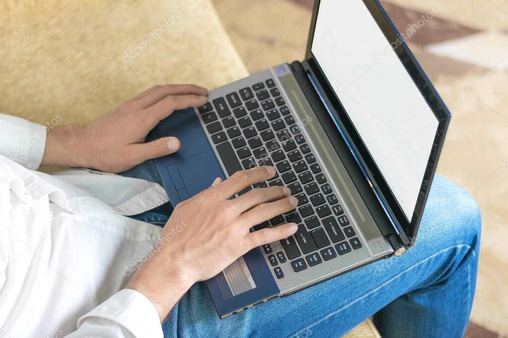young man in white shirt and blue jeans working on laptop, writes in notebook in home office sitting on cozy beige sofa Stay at home, online work, distance learning concept.