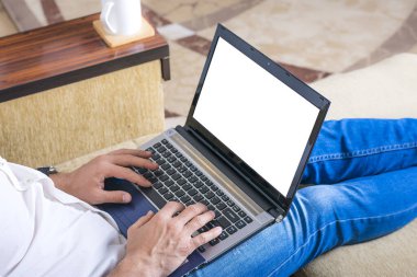 Beyaz gömlekli ve kot pantolonlu genç adam dizüstü bilgisayarda çalışıyor, ev bilgisayarında yazıyor rahat bej koltukta oturuyor evde oturuyor, online çalışıyor, uzaktan öğrenme konsepti geliştiriyor..