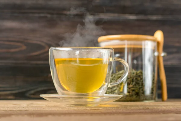 glass transparent cup of hot green tea with steam and can with tea leaves and spoon on wooden table Healthy drink, antistress beverage concept