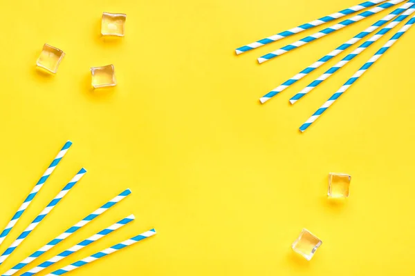 Drinking paper straws for party with blue stripes, ice cube on yellow background with copy space. Top view of colorful paper disposable eco - friendly straws for summer cocktails Flat lay.