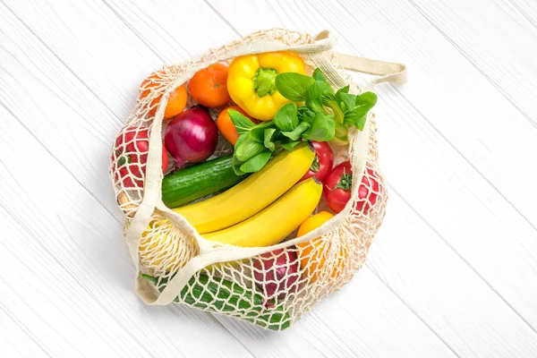 Full eco mesh bag of different health food - bell pepper, tomatoes, bananas, lemon, green, mandarin, cucumber, onions on white wooden background Top view