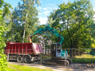 Şehrin avlusunda, metal bir çitin arkasındaki çimlerin ve ağaçların arasında bir turkuaz kazıcı bir yol yatağı kazıyor. Yakınlarda kırmızı bir damperli kamyon var. Rusya, St. Petersburg şehri, Ağustos 2019.