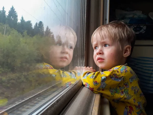 Un piccolo ragazzo biondo di tre anni cavalca su un treno, mangia caramelle e guarda fuori dalla finestra, dietro la quale vola un bellissimo paesaggio autunnale. . — Foto Stock