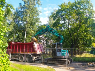 Şehrin avlusunda, metal bir çitin arkasındaki çimlerin ve ağaçların arasında bir turkuaz kazıcı bir yol yatağı kazıyor. Yakınlarda kırmızı bir damperli kamyon var. Rusya, St. Petersburg şehri, Ağustos 2019.