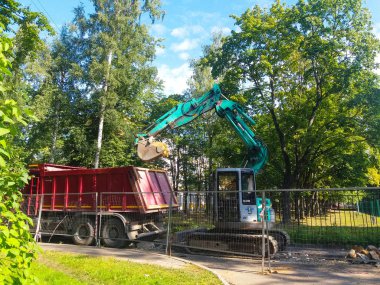 Şehrin avlusunda, metal bir çitin arkasındaki çimlerin ve ağaçların arasında bir turkuaz kazıcı bir yol yatağı kazıyor. Yakınlarda kırmızı bir damperli kamyon var. Rusya, St. Petersburg şehri, Ağustos 2019.