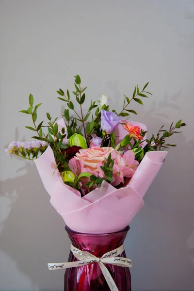 Un délicat bouquet de différentes fleurs dans un emballage rose clair se tient dans un vase bordeaux sur un fond gris . — Photo