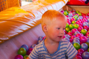 Oyun merkezinde üç yıllık sarışın bir çocuk renkli plastik toplarla havuzda oynuyor.