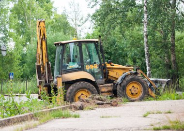 Fakir bir şehir. Yeşil ağaçların arka planında, bir patikayı tamir eden turuncu bir traktör görünür. İnşaat makineleri. Kazıcı.