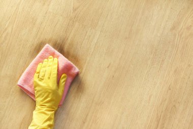 Sarı lastik eldivenli bir kadın eli, ahşap parke zeminindeki kiri pembe bir mikrofiber bezle siler. Metin için en üst görünüm kopyalama alanı.